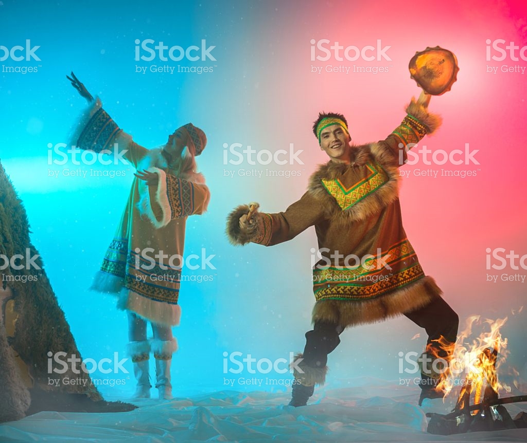 The beautiful young woman and young man with appearance of Siberian indigenous people are dressed in ethnic clothing.  The blonde young woman and attractive man are dancing a folk dance. The man is drumming in shaman drum. Theatrical performance in ethnic decoration, studio shooting in smoke